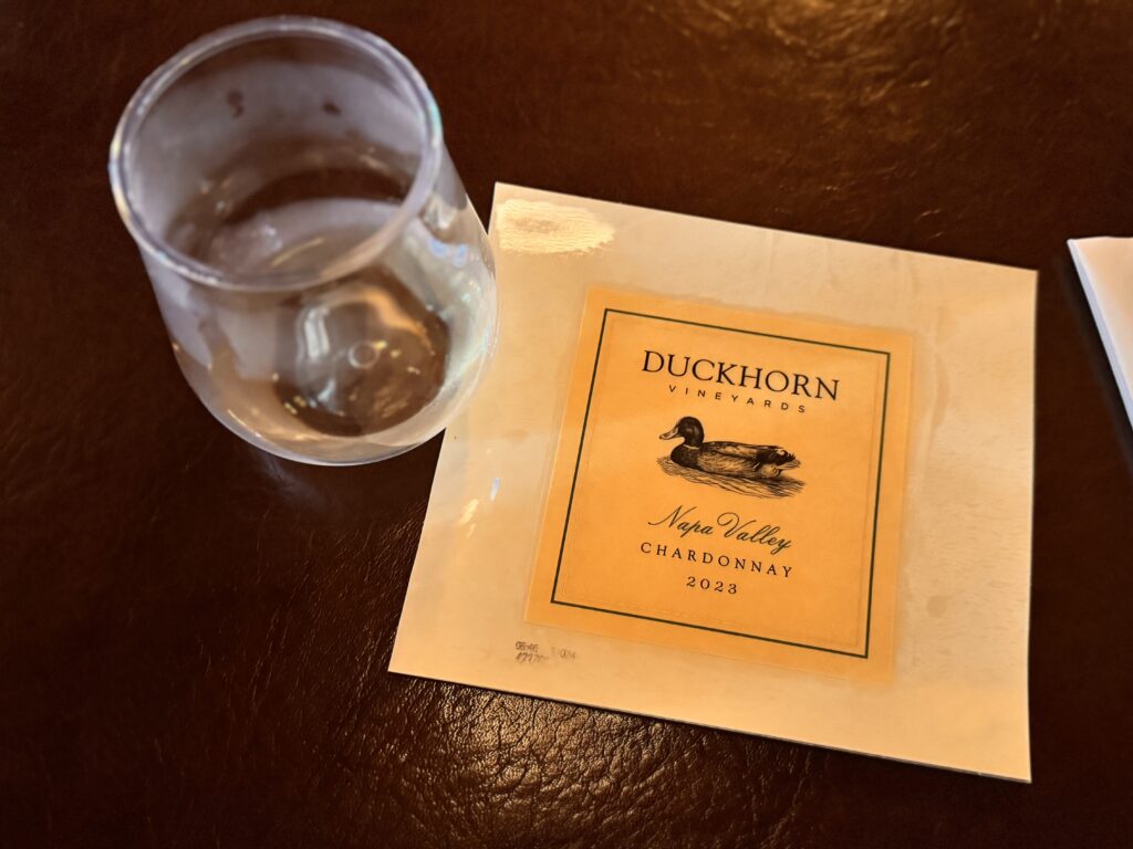 東京ディズニーシーのワインプログラムで提供されたDUCKHORN VINEYARDSのナパ・ヴァレー産シャルドネ2023年ヴィンテージ。白ワインのグラスとラベルがテーブルに置かれている。